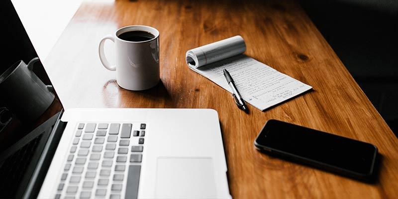 Wooden desk with laptop, iPhone, coffee mug, notepad, and pen